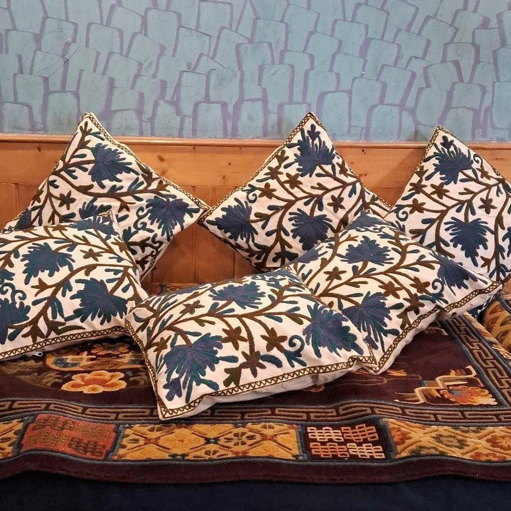 Decorative embroidered pillows with blue floral patterns on a traditional rug against a wooden panel and blue textured wall