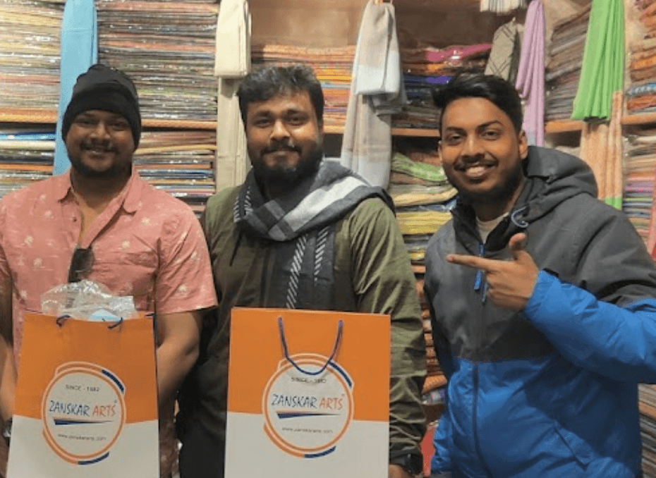 Three men smiling in a colorful textile shop holding orange Zanskar Arts shopping bags filled with traditional fabrics and shawls